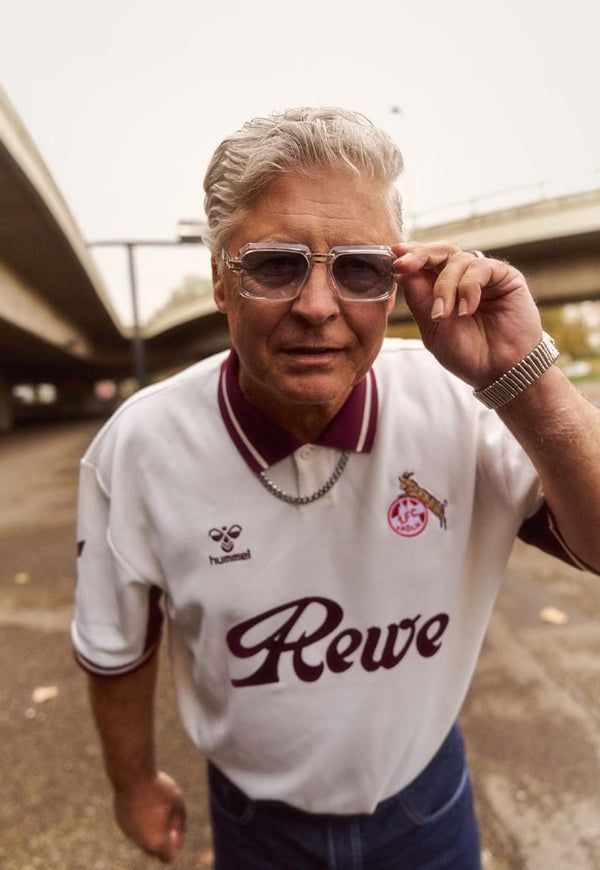 Man wearing a white sports jersey with 'Rewe' logo and sunglasses, standing outdoors.
