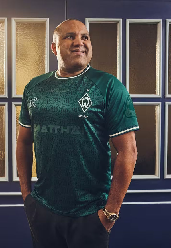 Ailton wearing a green anniversary shirt with visible branding in front of a decorative wall.